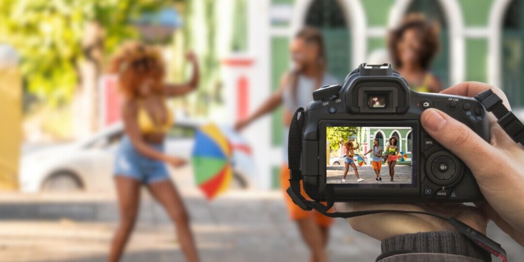 Fotografia do carnaval de rua em Salvador, Bahia. A imagem mostra duas mulheres e um homem fantasiados dançando e sorrindo em frente à câmera. Ao fundo uma imagem do pelourinho os foliões celebra a festa com música e alegria.